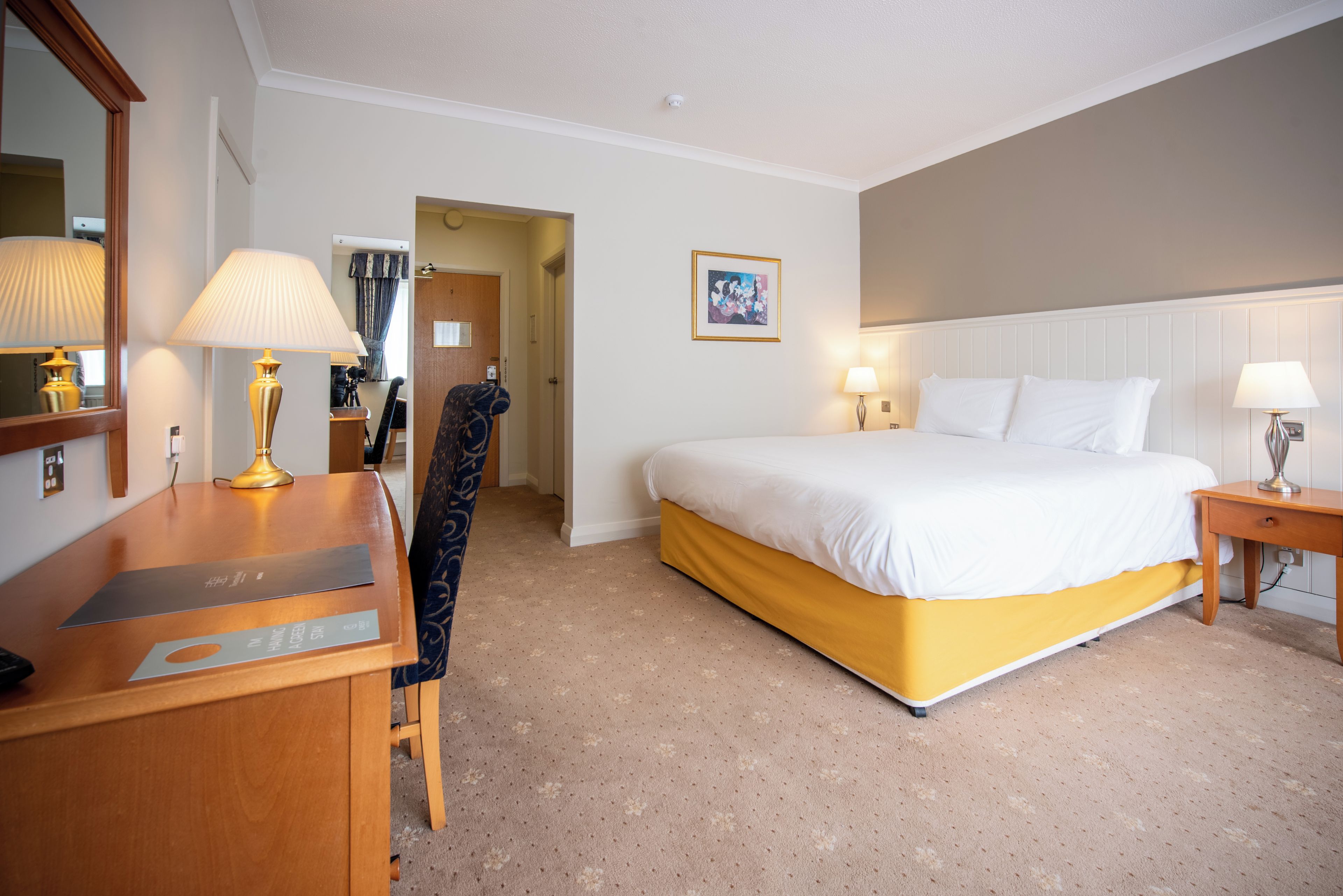 A hotel room with a bed, a desk, a chair, a lamp, and a painting on the wall.