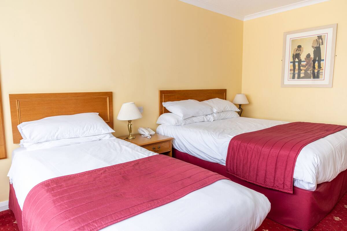 Ein Hotelzimmer mit zwei Betten, jeweils mit weißer Bettwäsche und einer roten Decke. An den Nachttischen stehen Lampen und ein Telefon.