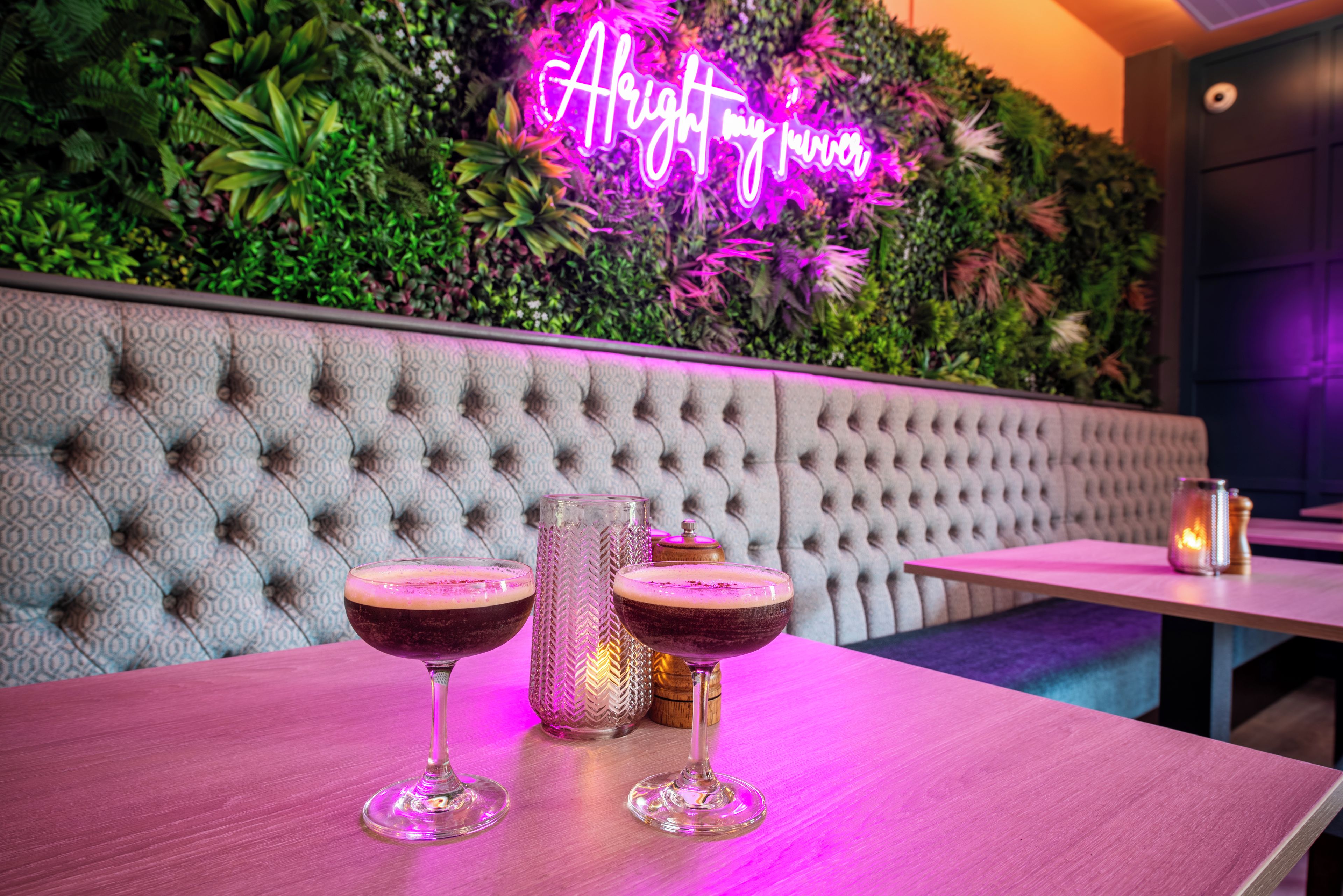 Zona de bar con un letrero de neón, dos vasos de una bebida oscura y un candelabro decorativo.