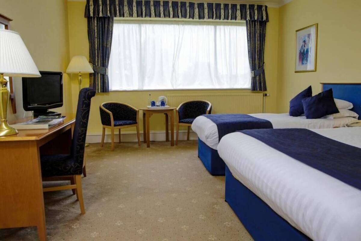 Ein Hotelzimmer mit zwei Betten, einem Schreibtisch mit Computer, einem Stuhl, einem kleinen Tisch und einem Fenster mit Vorhängen.