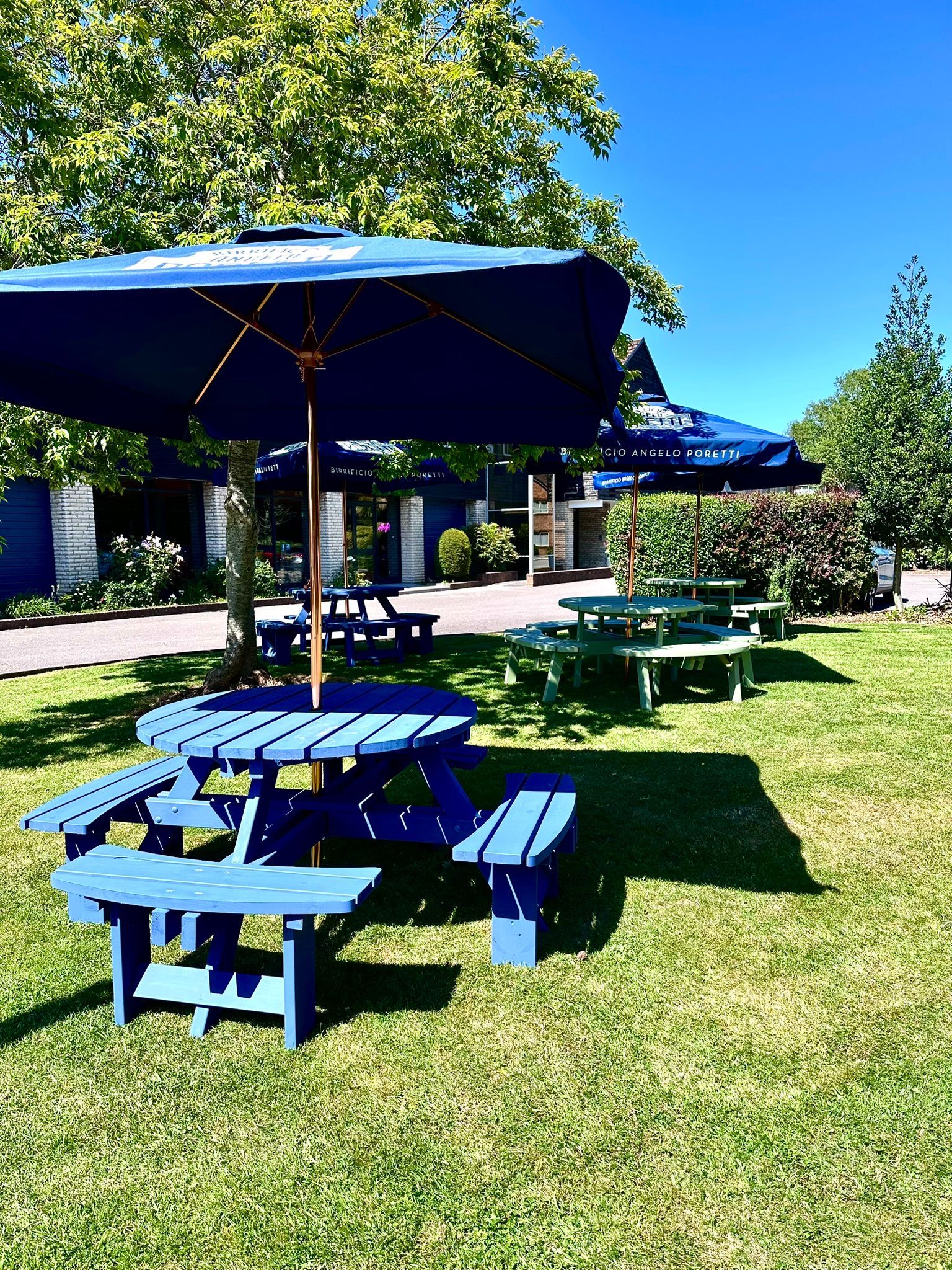 An outdoor seating area with blue picnic tables and umbrellas, a building with signage, and a clear blue sky.