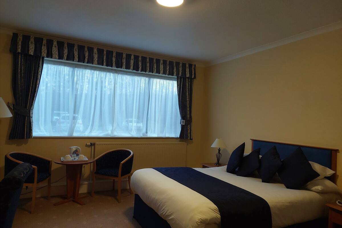 Ein Hotelzimmer mit einem Bett, zwei Stühlen, einem kleinen Tisch, einem Fenster mit Vorhängen und einem Nachttisch mit Lampe.