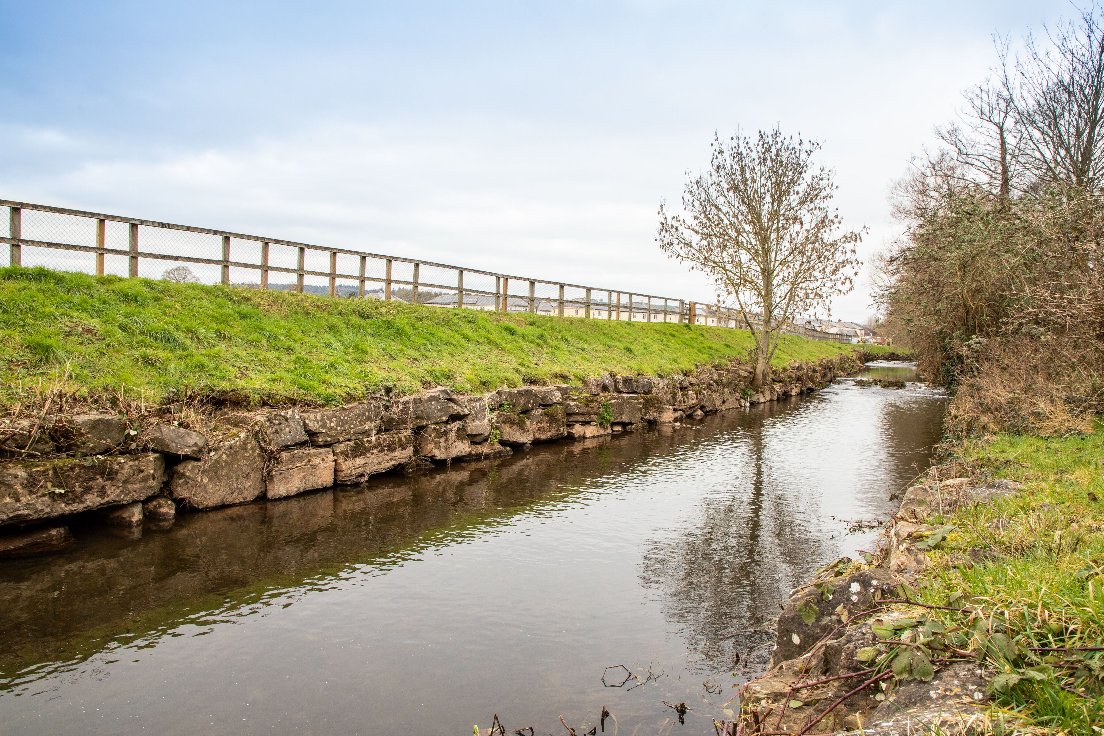 A narrow water channel with a stone wall on one side and a wooden fence on the other.