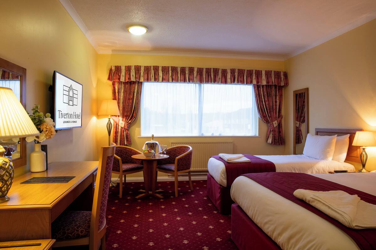 Ein Hotelzimmer mit zwei Betten, einem Schreibtisch mit Stuhl, einem Tisch mit Blumenvase und einem Fenster mit Vorhängen.