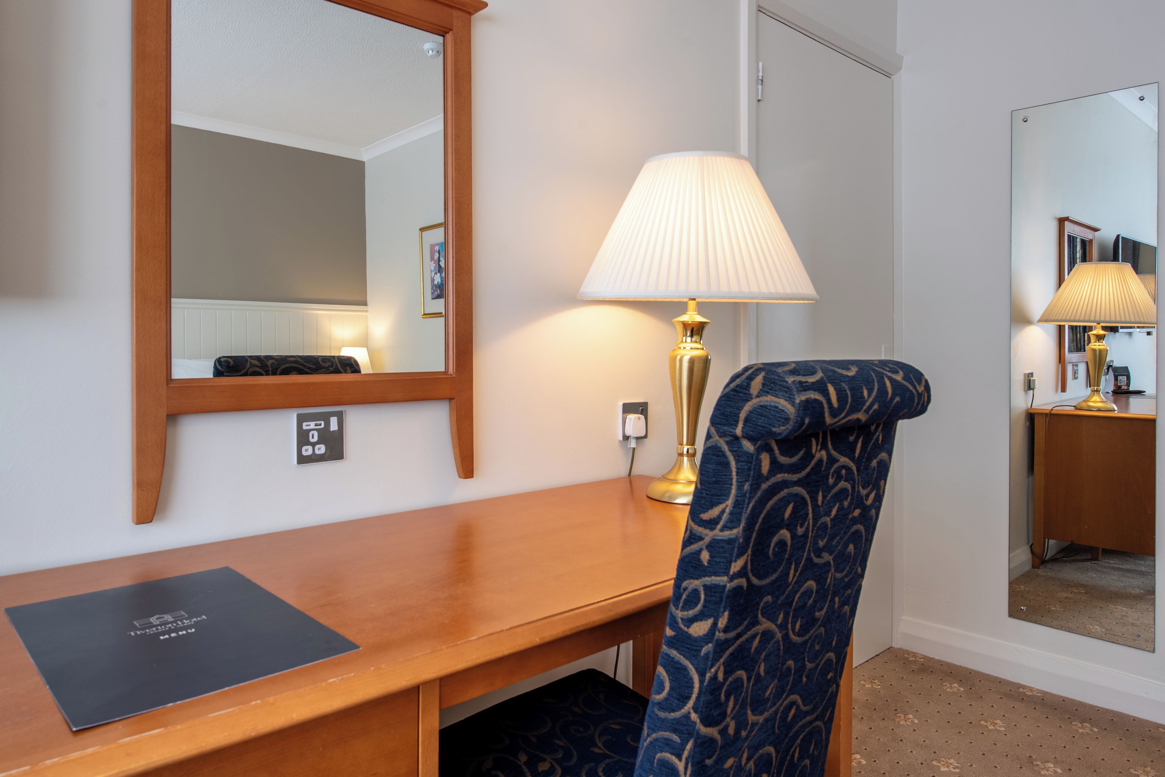 A hotel room with a wooden desk, a blue patterned chair, a wall-mounted mirror, and a bedside lamp.