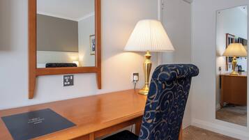 A hotel room with a wooden desk, a blue patterned chair, a wall-mounted mirror, and a bedside lamp.