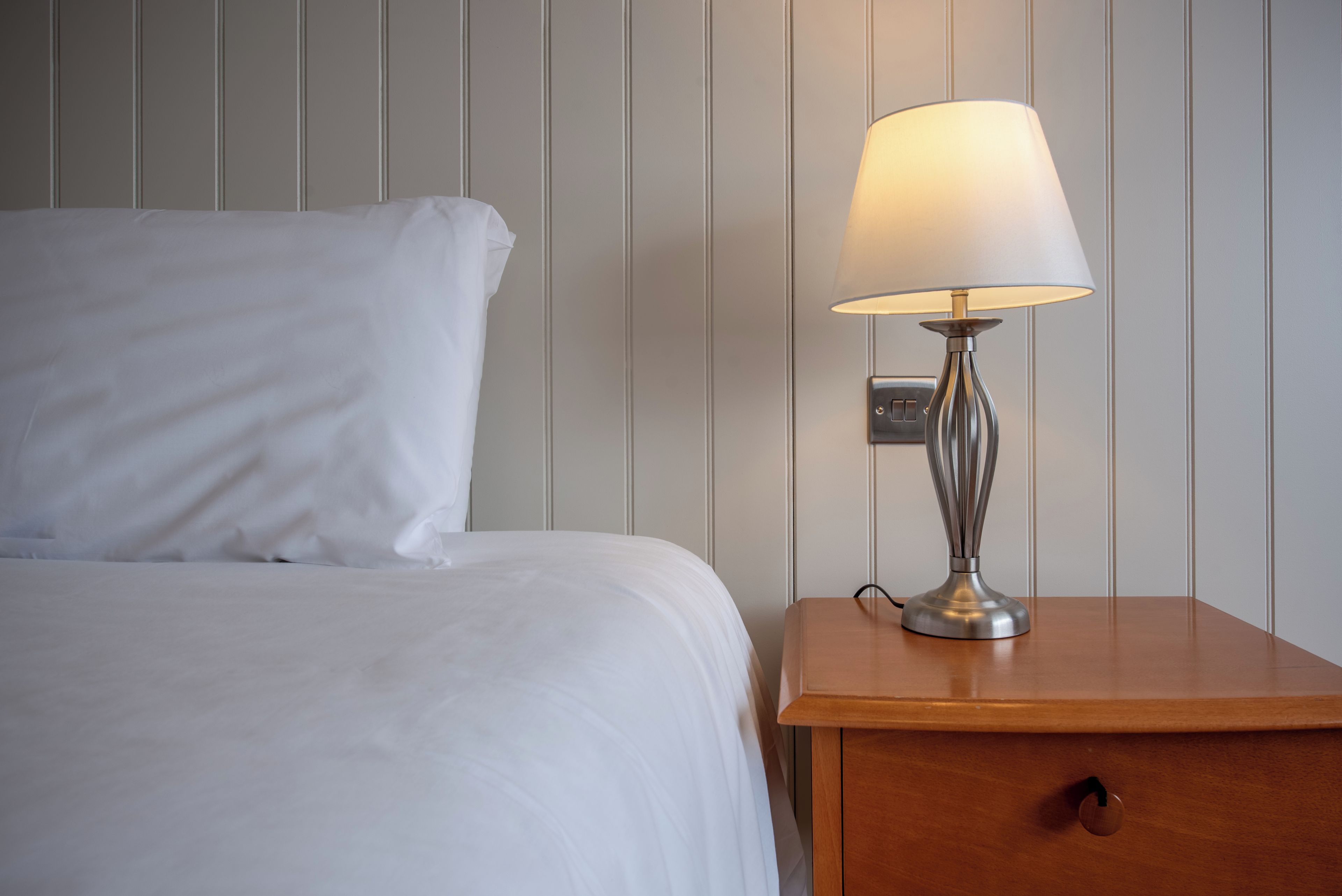 A neatly made bed with white linens, a bedside table with a lamp, and a wall with vertical paneling.