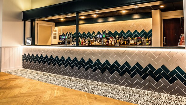 Een bar met een wand van patroon tegels, een houten vloer en diverse flessen achter de bar.