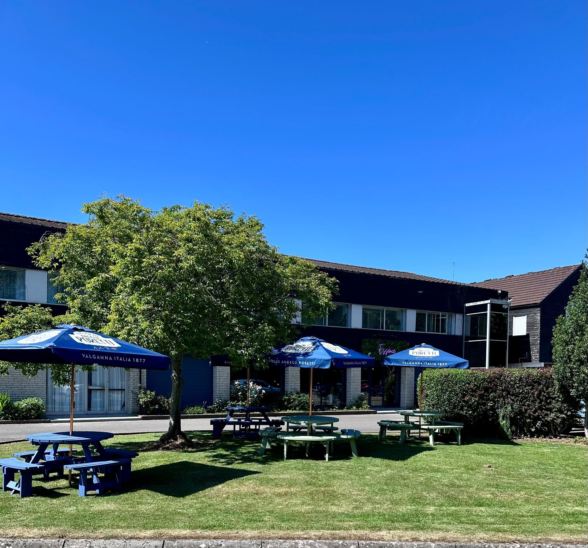An outdoor seating area with tables and blue umbrellas, a building with signage, and a tree in the foreground.
