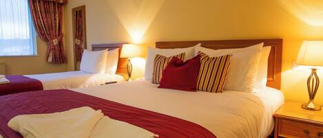 Une chambre d’hôtel avec deux lits, chacun doté d’une tête de lit en bois, de draps blancs et d’éléments décoratifs rouges. On y trouve des lampes de chevet et une table de chevet en bois avec un tiroir.