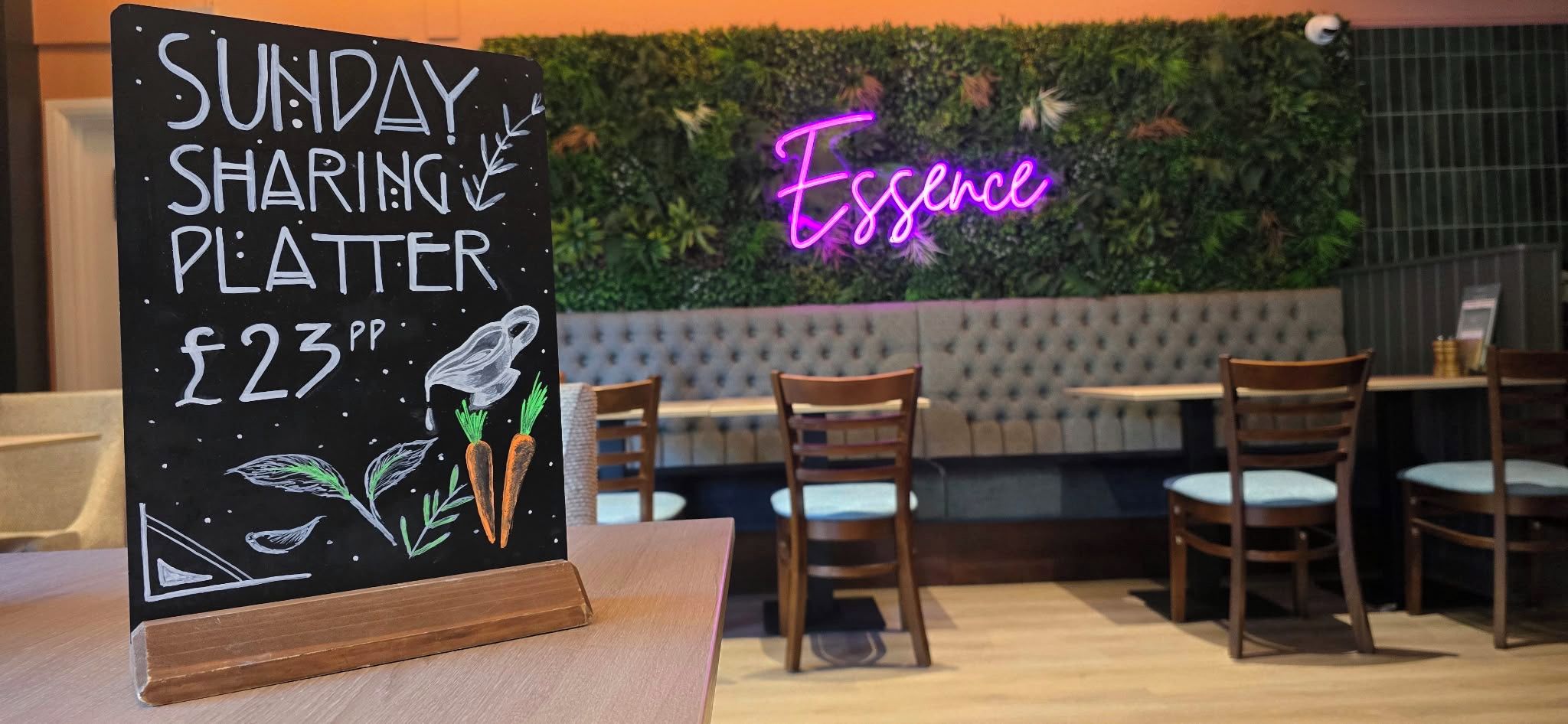 A sign for a Sunday sharing platter priced at £23, a neon sign reading 'Essence,' and a seating area with wooden chairs and a cushioned bench.