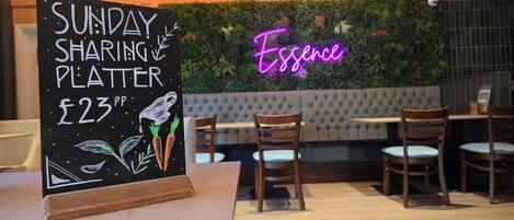 A sign for a Sunday sharing platter priced at £23, a neon sign reading 'Essence,' and a seating area with wooden chairs and a cushioned bench.