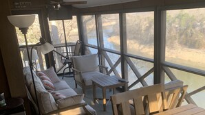 Ein sonnendurchflutetes Zimmer mit einem Holztisch, Stühlen und einer Couch. Große Fenster bieten Blick auf ein Gewässer und Bäume.