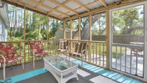 Une véranda couverte, meublée de meubles en osier et dotée d’une table en verre, donnant sur un jardin avec des arbres et une clôture en bois.