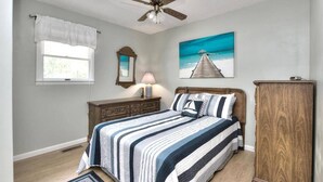 Une chambre à coucher comprenant un lit, un ventilateur de plafond, une fenêtre avec des rideaux, une commode, une lampe et un tableau représentant une jetée.