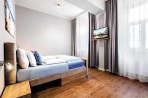 Quarto moderno com cama de madeira, televisão montada e cortinas leves.