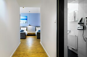 Corridoio di un hotel con pavimento in legno, due letti con lenzuola blu, un quadro appeso al muro e un termosifone nel bagno.