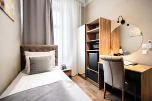 Quarto de hotel com cama, escrivaninha, cadeira, guarda-roupa, espelho e janela com cortinas.