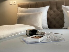 Uma cama bem arrumada com travesseiro branco, cabeceira marrom e uma taça de vinho de vidro transparente sobre uma bandeja com uma caixa de chocolates.