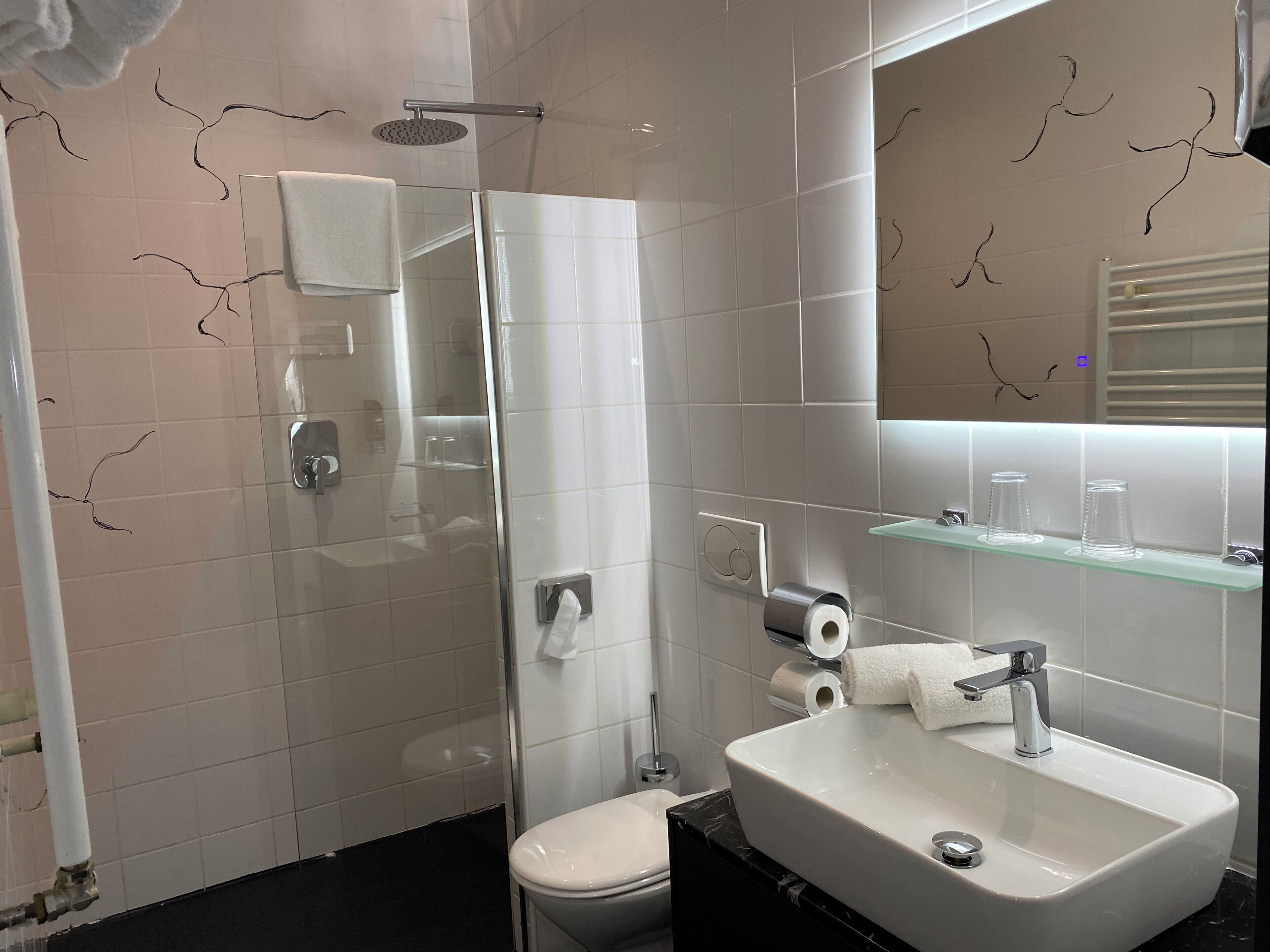 A modern bathroom with a glass shower enclosure, a white toilet, a sink with a mirror above it, and a glass shelf with toiletries.