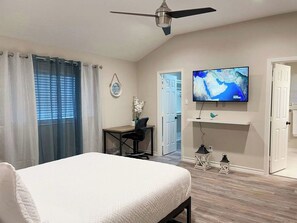 Un dormitorio con cama, escritorio con silla, televisión en la pared y ventilador de techo.