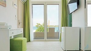 Habitació amb una porta corredissa de vidre que dona a un balcó, un refrigerador blanc, una taula petita amb una làmpada i un televisor fixat a la paret.
