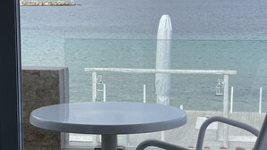 Un balcó amb una taula rodona i una cadira blava, amb vista a un mar tranquil i a una costa rocosa.