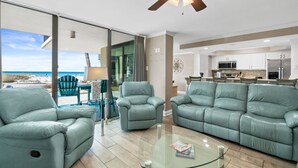 Un soggiorno moderno con un divano, poltrone e un tavolino in vetro. Si nota una ventola a soffitto, vista sulla spiaggia e una cucina sullo sfondo.