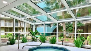 Zona de piscina con cerramiento de vidrio, que incluye un jacuzzi, rodeada de plantas y con vistas a un edificio con balcones.