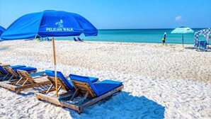 A beach with blue umbrellas labeled "PELICAN WALK," wooden lounge chairs, and a clear view of the ocean.