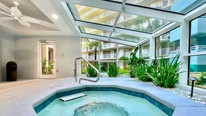 Piscina interior moderna con techo de cristal, rodeada de plantas tropicales y vista a un edificio residencial.