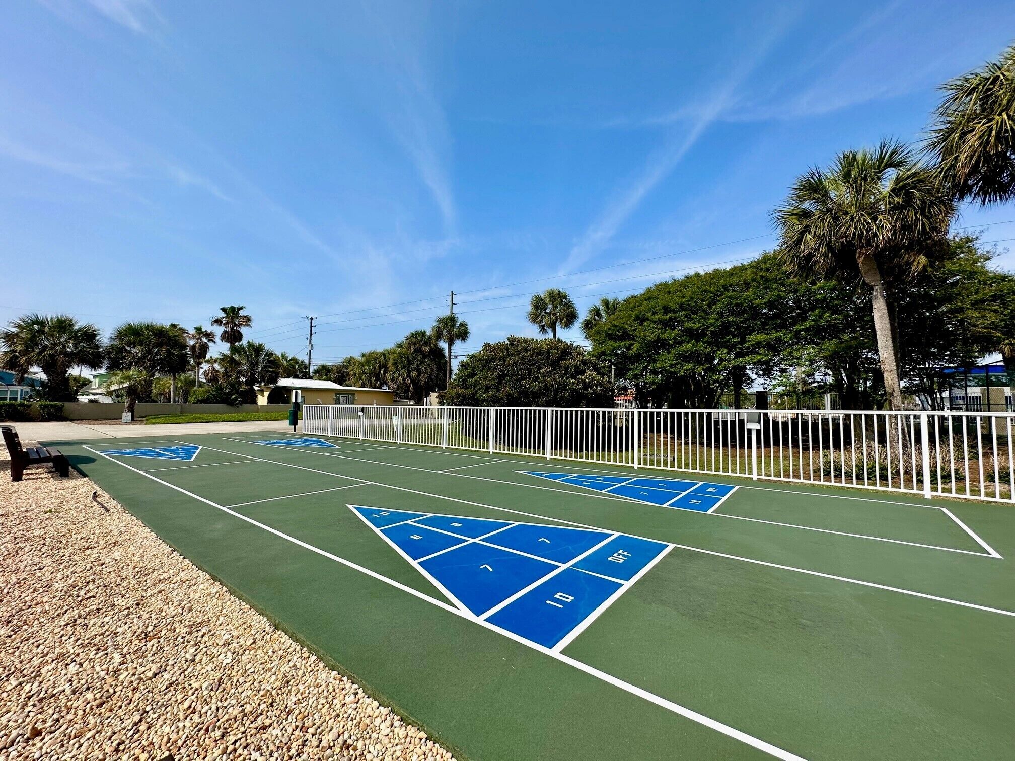 Ein Tennisplatz mit blauen Behindertenflächen, umgeben von einem weißen Zaun und Palmen.
