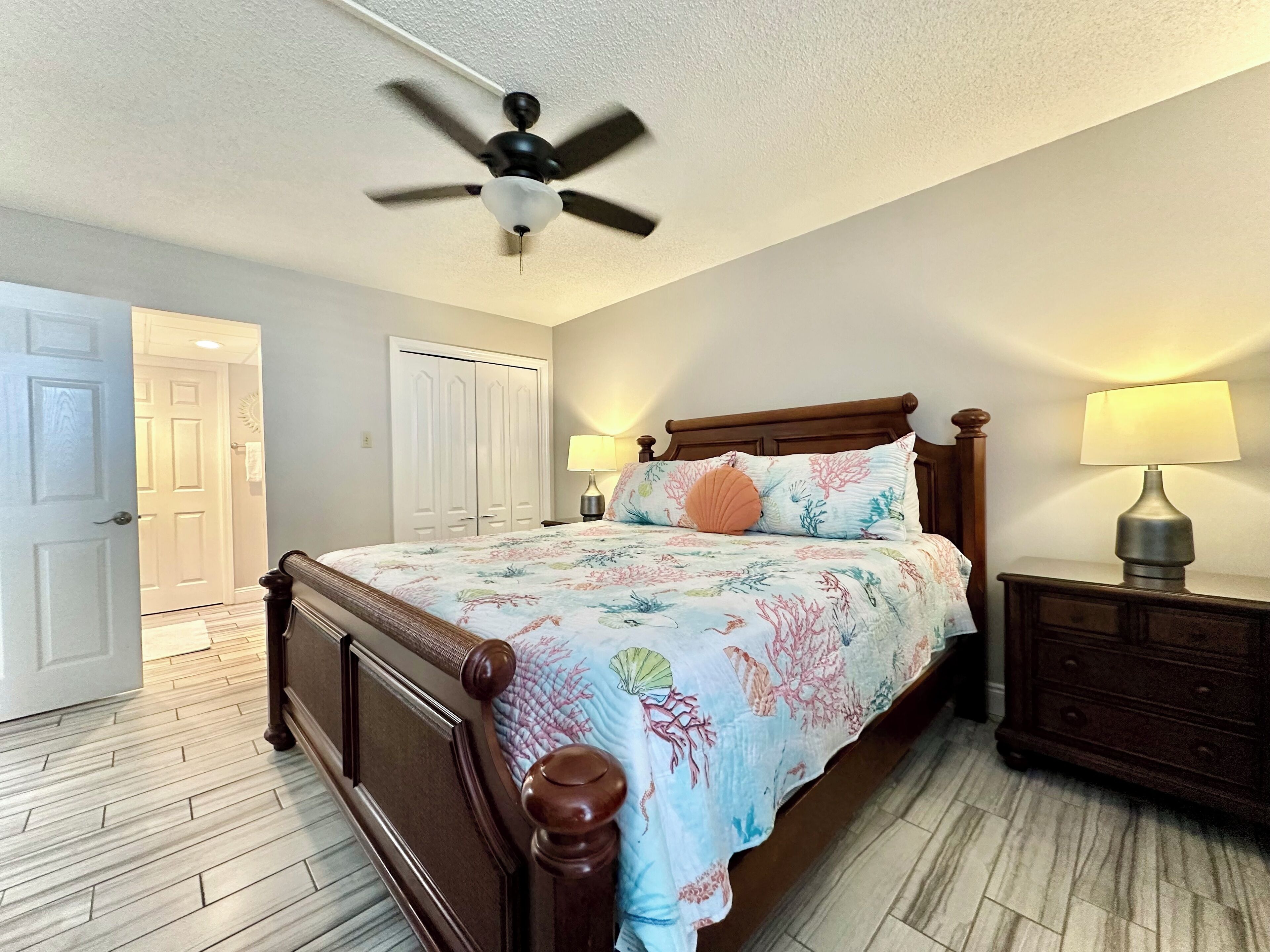 A bedroom with a wooden bed, two bedside lamps, a ceiling fan, and a door leading to another room.