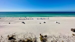 A beach with white sand and a clear blue sky. There are lounge chairs and umbrellas along the shoreline.