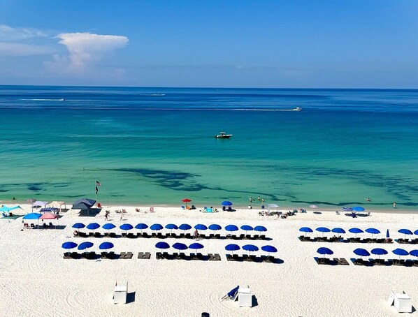 A beach with rows of blue umbrellas and sun loungers, a clear blue sea, and a few boats in the distance.