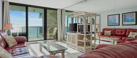 Un salón amplio con un sofá de cuero rojo, una mesa de centro de vidrio y un televisor sobre un mueble blanco. Hay un balcón con vista al mar.