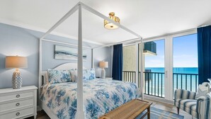 A four-poster bed with a blue floral bedspread, a wooden bench, a white nightstand with a patterned lamp, and a view of the ocean through large windows.