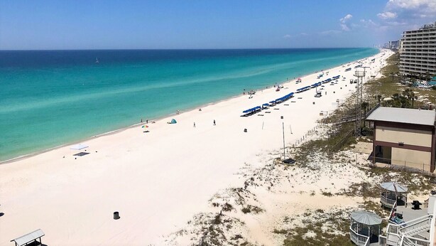 A beach with a long stretch of umbrellas and sun loungers, a clear blue sky, and a building with multiple balconies overlooking the beach.