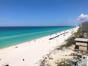 A beach with a long stretch of umbrellas and sun loungers, a clear blue sky, and a building with multiple balconies overlooking the beach. - Pet Friendly Beach Front Condo! FREE Beach Chairs! (Panama City Beach)