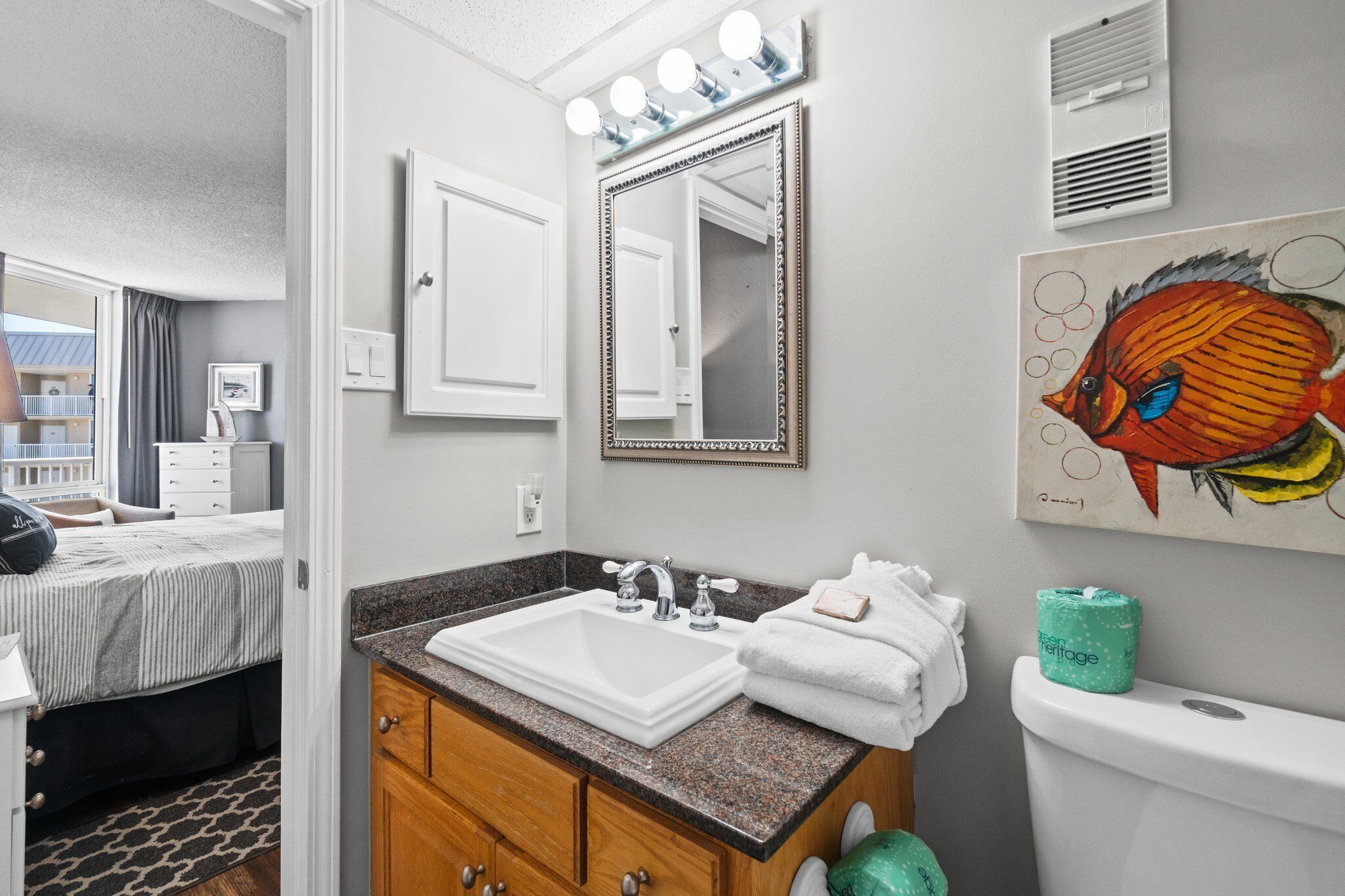 A bathroom with a sink, mirror, and toilet. There is a painting on the wall and a towel rack.