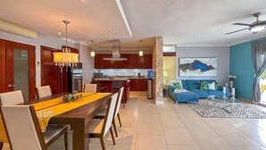 A modern living room with a blue sofa, a dining area with a wooden table, and a kitchen with dark wooden cabinets. - Incredible 3 BR Penthouse w BBQ & Jacuzzi (Punta Cana)