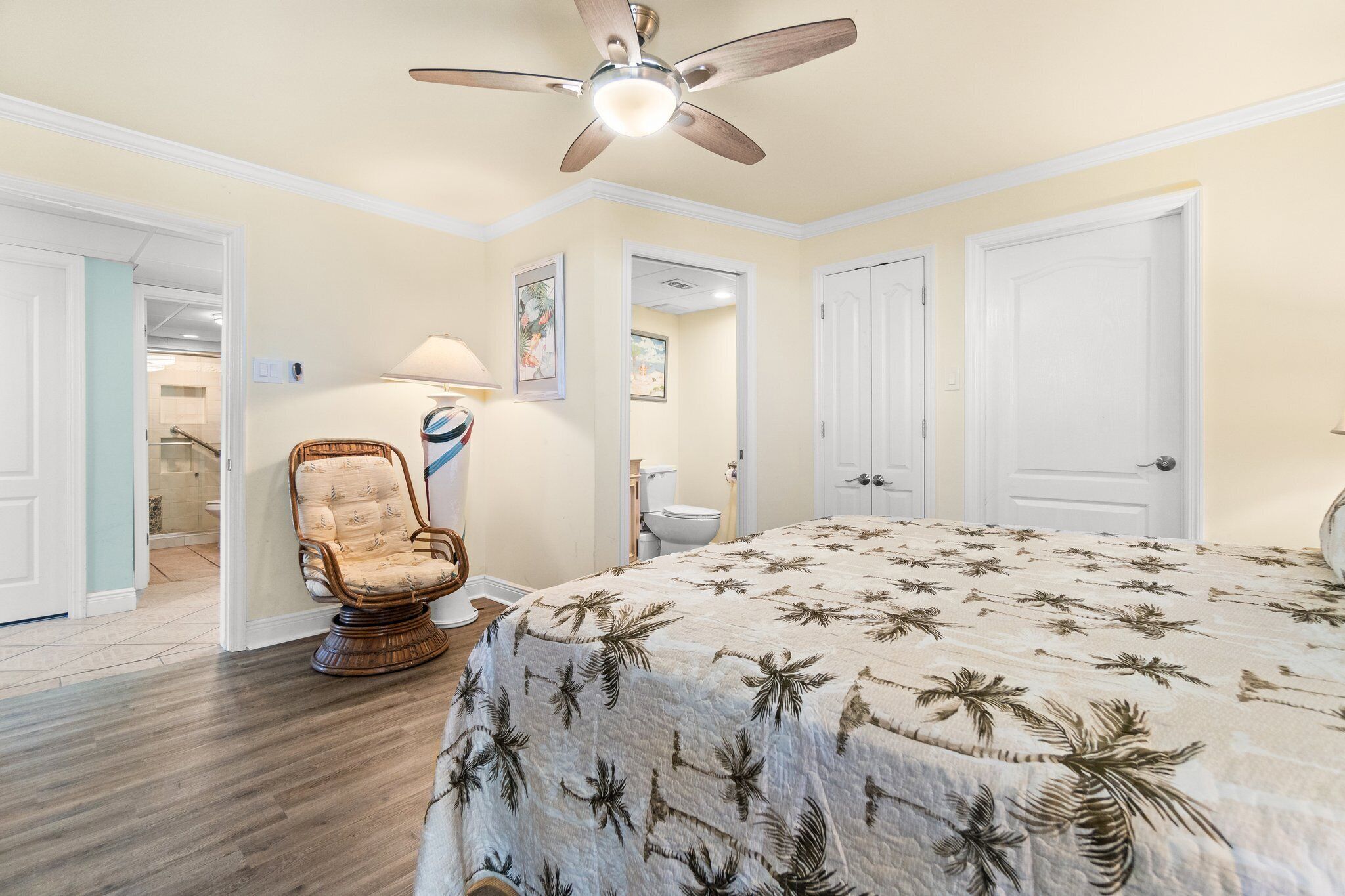 Une chambre à coucher comprenant un lit, un ventilateur de plafond, une chaise et une porte donnant sur une salle de bain.