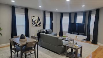 A modern living room with a grey sofa, a dining table with chairs, a TV, and a painting on the wall.