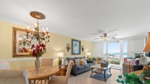 Ein geräumiges Wohnzimmer mit einer Chandelier, einem Sofa, einem Esstisch und Blick auf den Strand durch große Fenster.