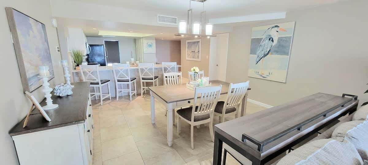 A dining area with a wooden table and white chairs, a kitchen in the background, and a painting of a bird on the wall.