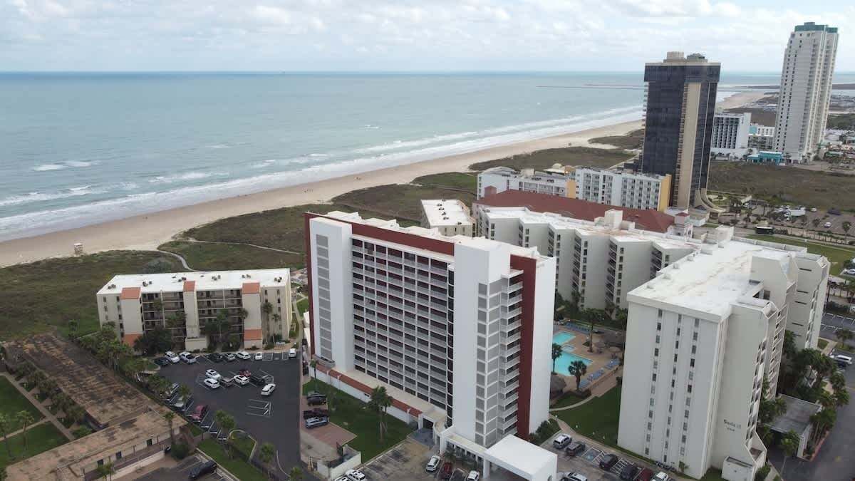 A coastal hotel with multiple buildings, a parking lot, and a beach view.