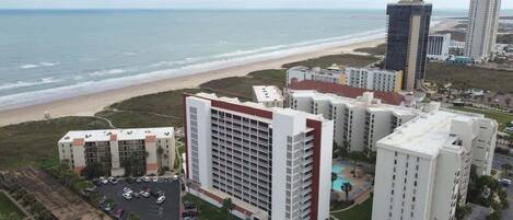 A coastal hotel with multiple buildings, a parking lot, and a beach view.