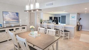 A modern kitchen with white cabinetry, a dining area with a wooden table and white chairs, and a view of the ocean. - Saida 3601 3 Bedroom Home by RedAwning (South Padre Island)