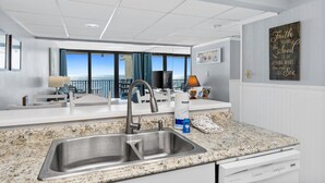 Cozinha moderna com pia de aço inoxidável, bancada de granito e vista para o oceano através de grandes janelas.