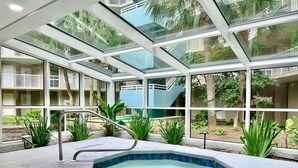 Uma área de piscina interior moderna com paredes de vidro, uma banheira de hidromassagem e vegetação ao redor.